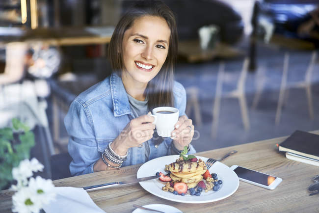 Ritratto di giovane donna sorridente che si gode frittelle e caffè nel caffè — Foto stock