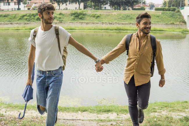 Портрет молодої пари-гея з рюкзаками, що йдуть рукою в руці на березі річки — стокове фото