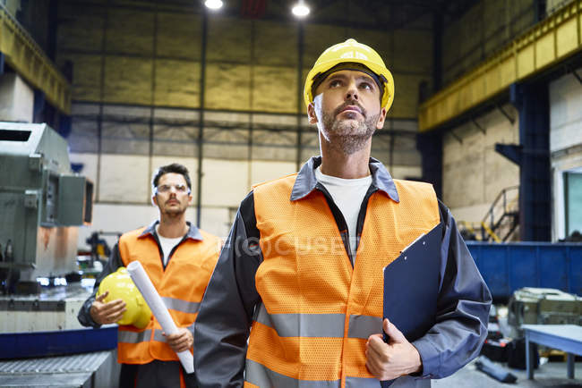 Hombres que usan ropa de trabajo protectora mirando alrededor en fábrica - foto de stock