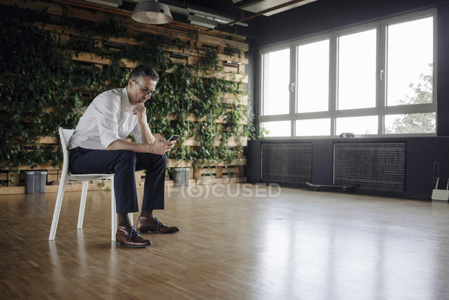 Бізнесмен сидить на кріслі в зеленому офісі дивлячись на стільниковий телефон — стокове фото