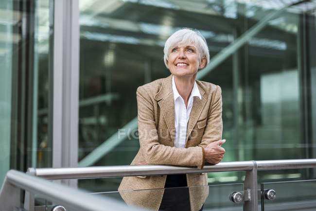 Портрет улыбающейся старшей бизнес-леди, опирающейся на перила в городе и смотрящей вверх — стоковое фото