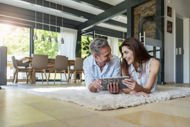 Coppia felice sdraiata sul tappeto a casa condividendo un tablet — Foto stock