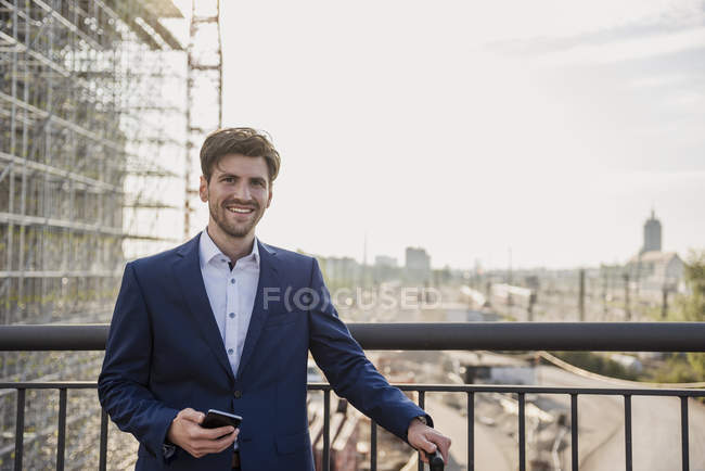Портрет усміхненого бізнесмена, що стоїть на мосту в місті, тримає мобільний телефон — стокове фото