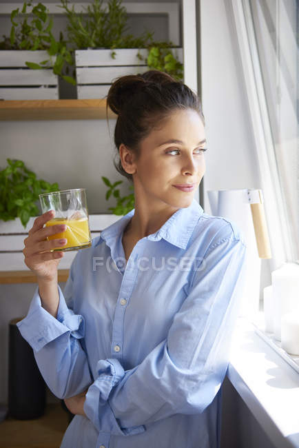 Giovane donna che tiene il bicchiere con succo d'arancia in cucina e sorride guardando la finestra — Foto stock