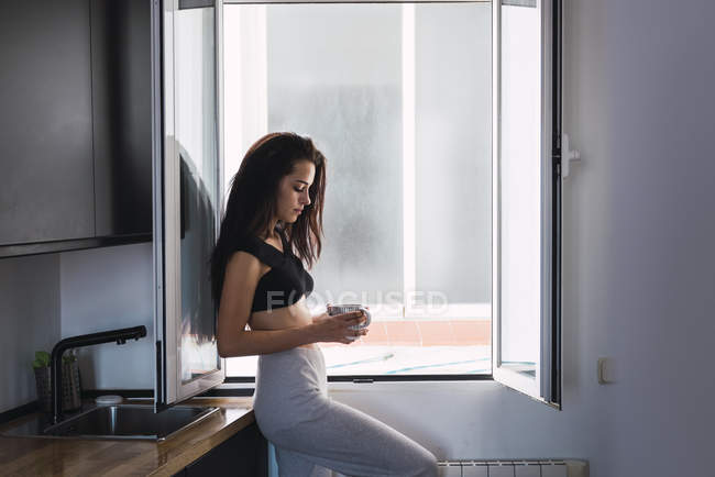 Серьёзная молодая женщина с чашкой кофе, мечтающая у окна дома — стоковое фото