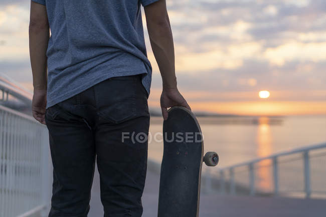 Молодой человек со скейтбордом на пляже на рассвете, вид сзади — стоковое фото