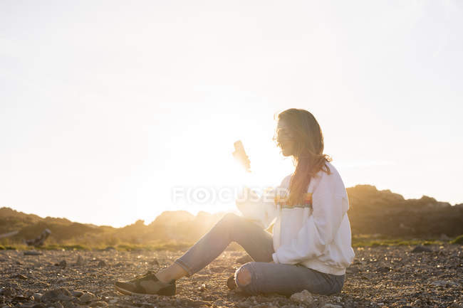 Young woman at the beach, using smartphone at sunset — Stock Photo