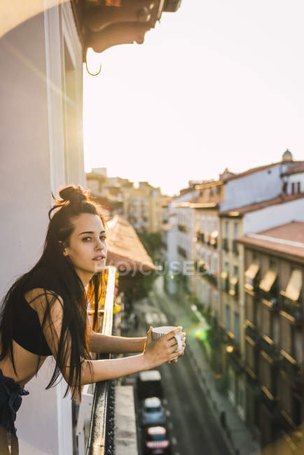 Приваблива молода жінка, що стоїть на балконі над містом на заході сонця з чашкою кави — стокове фото