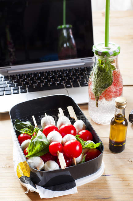 Lunch box of Caprese salad, bottle of infused water and laptop on desk — Stock Photo