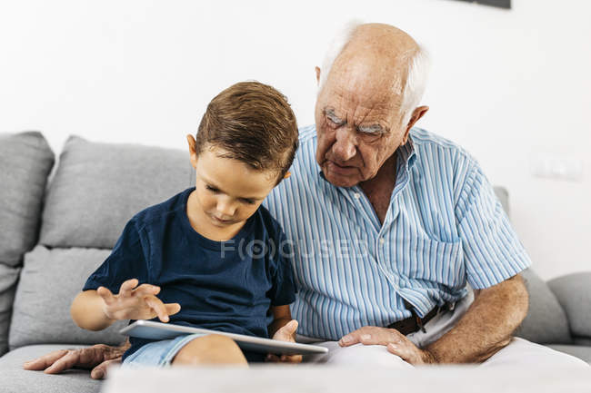 Abuelo y nieto sentados juntos en el sofá en casa y usando tableta digital - foto de stock