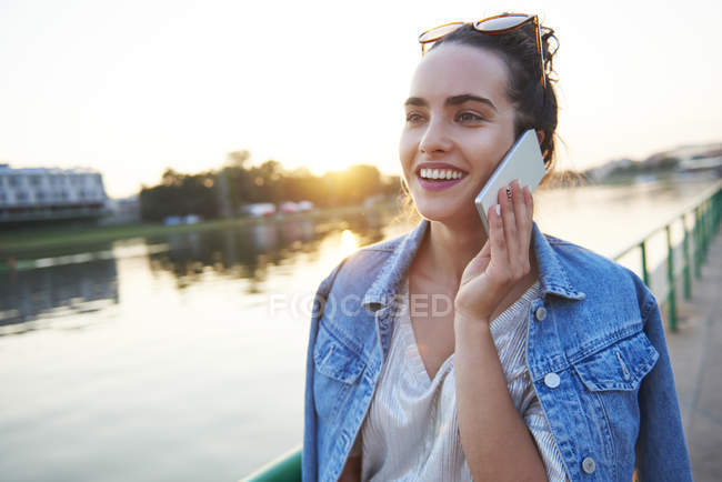 Усміхнена молода жінка на мобільному телефоні біля річки на заході сонця — стокове фото