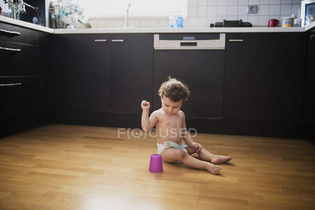 Bambino seduto sul pavimento in cucina a giocare con la tazza di plastica — Foto stock