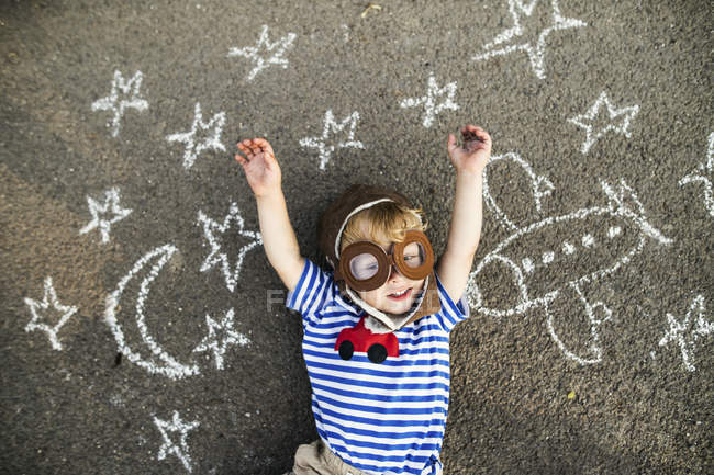Retrato de un niño sonriente con sombrero de piloto y gafas, acostado sobre asfalto pintado con avión, luna y estrellas - foto de stock