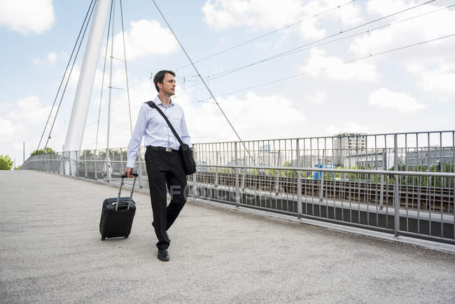 Uomo d'affari in città con valigia rotolante che attraversa un ponte — Foto stock