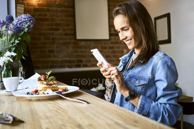 Smiling young woman with plate of pancakes using phone in cafe — Stock Photo