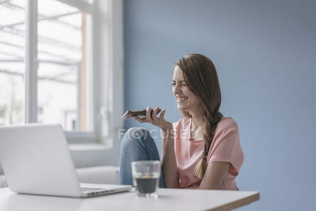 Donna ridente a casa seduta alla scrivania con computer portatile e utilizzando smartphone — Foto stock