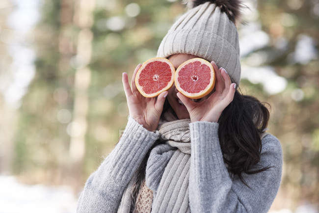 Mujer joven que ríe vistiendo prendas de punto que cubren sus ojos con mitades de pomelo - foto de stock