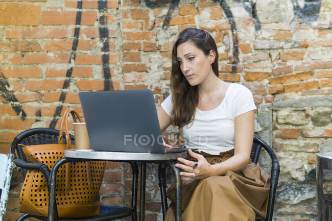 Mujer usando el ordenador portátil en la cafetería al aire libre, sentado en la mesa en la silla, pared de ladrillo - foto de stock