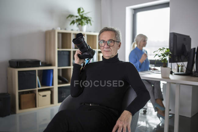 Hombre mayor con cámara sentado en la oficina con un colega trabajando detrás de él - foto de stock