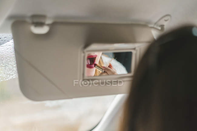 Mujer espejada en espejo retrovisor aplicando lápiz labial rojo en el coche - foto de stock