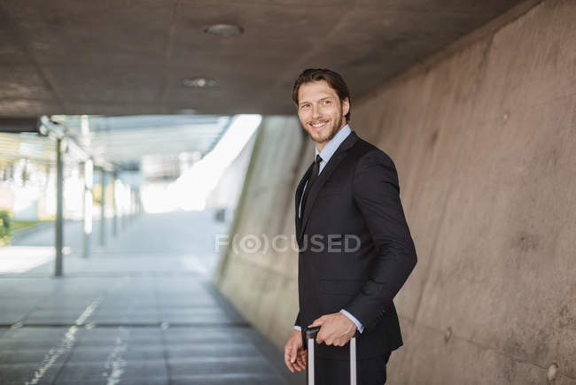 Hombre de negocios sonriente con maleta rodante de pie en el paso subterráneo - foto de stock