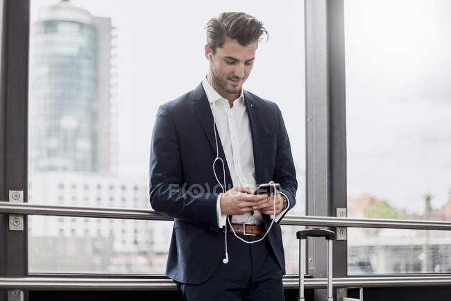 Молодой бизнесмен у окна с сотовым телефоном, наушниками и чемоданом — стоковое фото