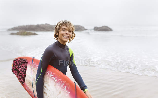 Spain, Aviles, portrait of smiling young surfer carrying surfboard on the beach — Stock Photo