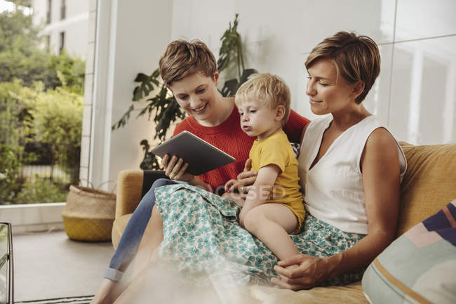 Dos madres leyendo un libro de tableta con su hijo en casa - foto de stock