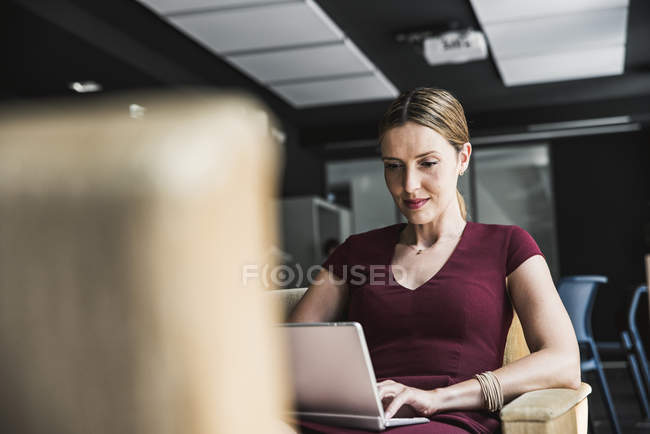 Businesswoman in office lounge wearing burgundy dress using laptop — Stock Photo