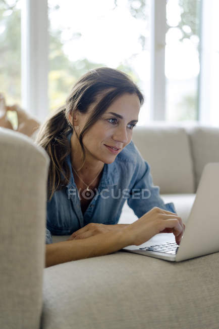 Sorridente donna matura sdraiata sul divano a casa utilizzando il computer portatile — Foto stock