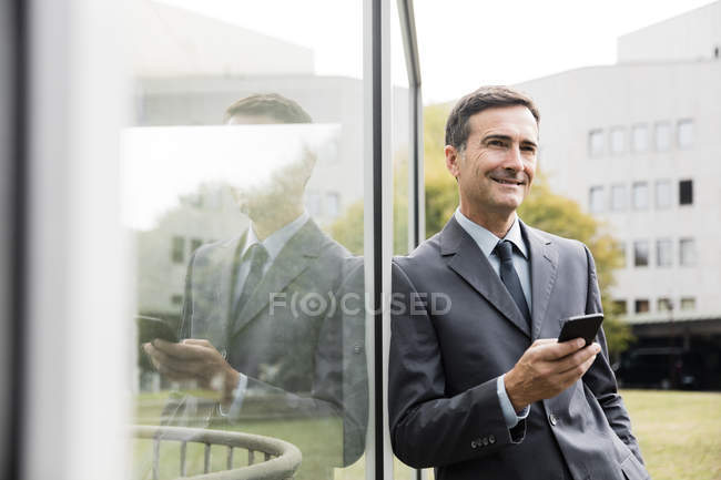 Усміхнений бізнесмен з мобільним телефоном, що спирається на скляний фронт — стокове фото