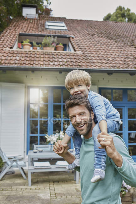 Portrait of happy father carrying son on shoulders in front of their home — Stock Photo