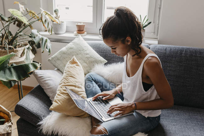 Mujer joven sentada en el sofá en casa usando el ordenador portátil - foto de stock