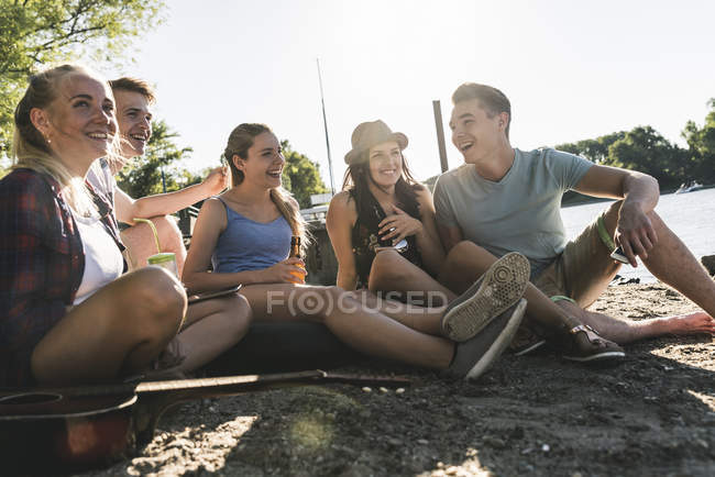 Gruppe fröhlicher Freunde sitzt mit Getränken am Flussufer — Stockfoto