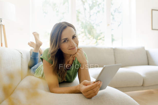 Портрет улыбающейся зрелой женщины, лежащей дома на диване и использующей планшет — стоковое фото