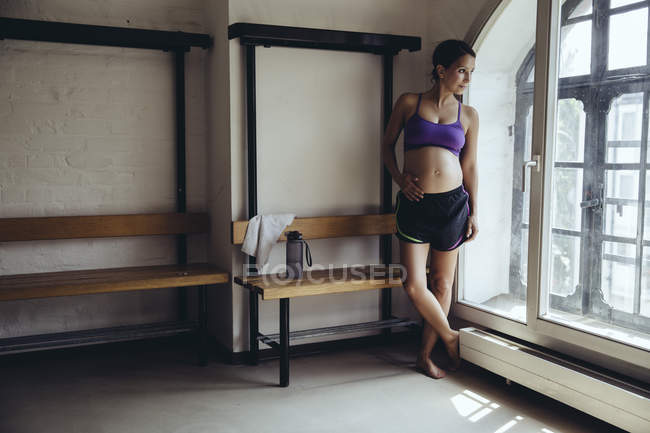 Deportiva mujer embarazada de pie en vestuario mirando por la ventana - foto de stock