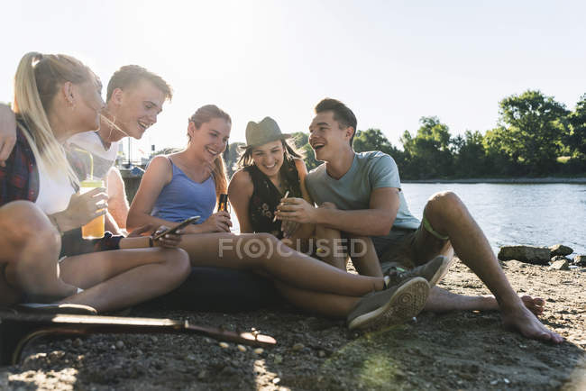 Grupo de amigos felizes sentados à beira do rio e brindando com bebidas — Fotografia de Stock
