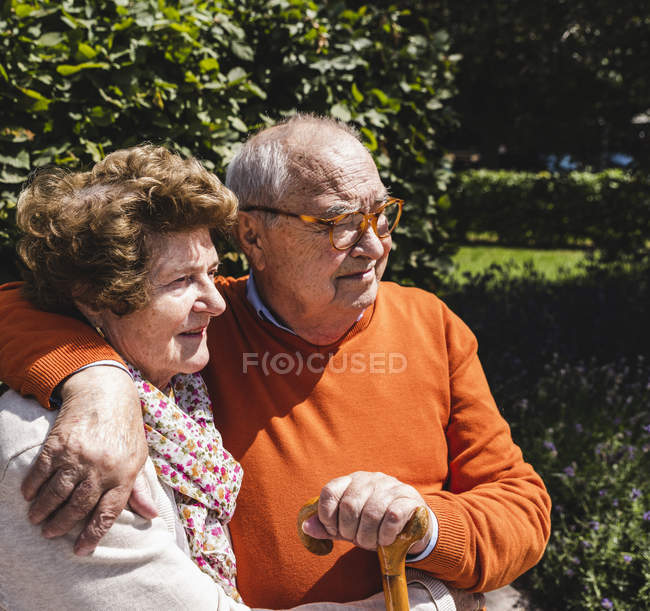 Couple âgé assis sur un banc dans le parc avec les bras autour — Photo de stock