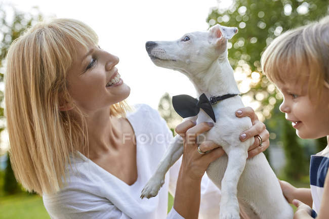 Mujer feliz con hija sosteniendo perro usando una corbata al aire libre - foto de stock