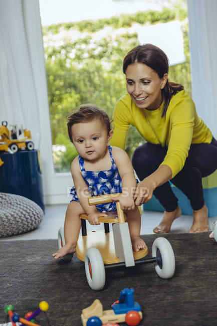 Happy mother with baby daughter driving toy car in living room — Stock Photo