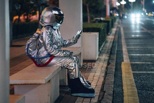 Homme de l'espace assis sur le banc à l'arrêt de bus la nuit et tenant son téléphone portable — Photo de stock
