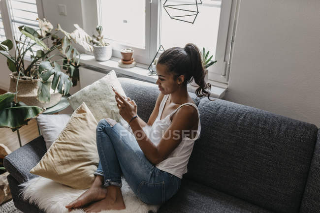 Молодая женщина сидит дома на диване с помощью мобильного телефона — стоковое фото