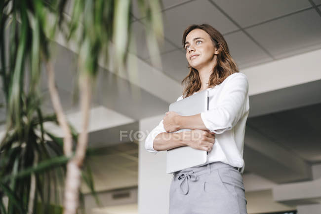 Businesswoman standing in office and holding laptop — Stock Photo