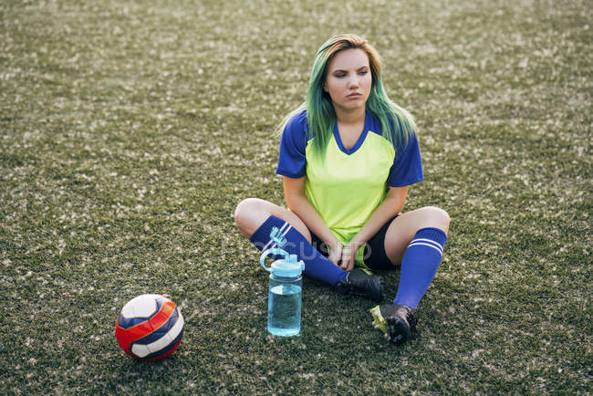 Jeune femme assise sur le terrain de football avec bouteille d'eau et ballon — Photo de stock