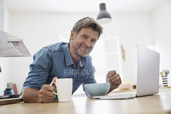 Porträt eines reifen Mannes in der Küche beim Frühstück, während er Laptop checkt — Stockfoto