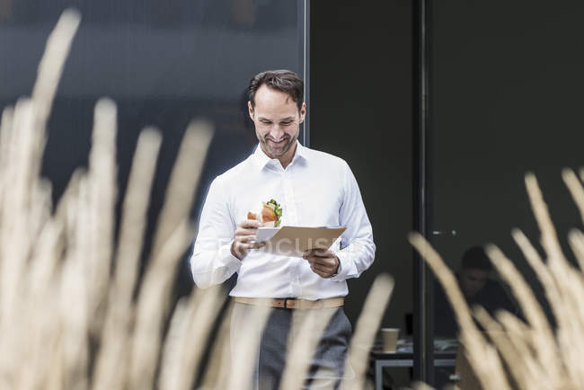 Un hombre de negocios sonriente con documentos de lectura de sándwiches frente al edificio de oficinas - foto de stock