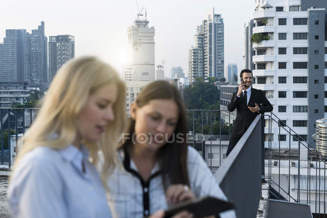 Бизнесмен говорит на смартфоне, в то время как женщины-коллеги ищут планшет на крыше города — стоковое фото