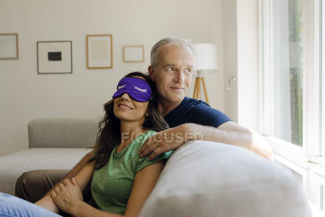 Sorridente coppia matura seduta sul divano a casa con la donna che indossa maschera per gli occhi — Foto stock