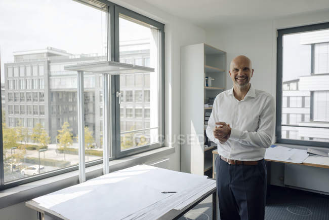 Homme d'affaires souriant debout dans son bureau à table avec un plan de construction avec les bras serrés — Photo de stock