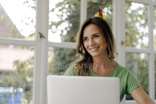 Lächelnde reife Frau am Fenster mit Laptop — Stockfoto